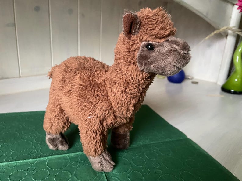 May include: A brown plush llama toy with a fluffy body, standing on a green surface. The llama has a detailed face with dark eyes and a soft, textured appearance. The toy is a cute and cuddly item.