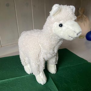 May include: A white plush llama toy with a fluffy texture. The soft toy has black eyes and stands on a green surface. The llama is a cute and cuddly toy.
