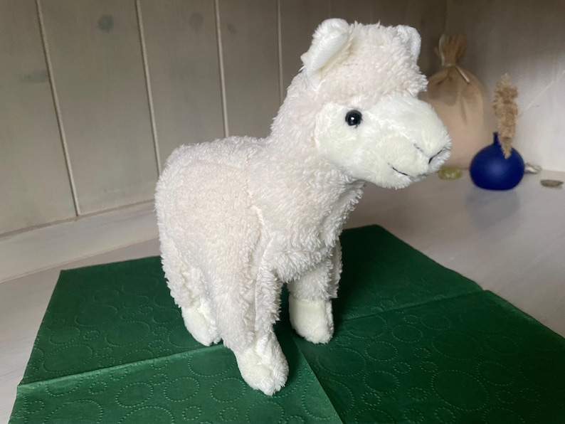 May include: A white plush llama toy with a fluffy texture. The soft toy has black eyes and stands on a green surface. The llama is a cute and cuddly toy.
