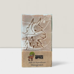 May include: A rectangular bar of soap with a carved alpaca design. The soap is wrapped in clear plastic and has a brown label with the text "HEUBERS ALPAKA" and "Honigmilch". The soap is light brown in color.