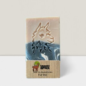 May include: A rectangular bar of soap with a carved alpaca design. The soap has a light beige top, a blue and white marbled center, and a brown label with text and a logo. The label reads "HEUBERS AIPAKA" and "Seife mit Alpakakeratin F&uuml;r Ihn."