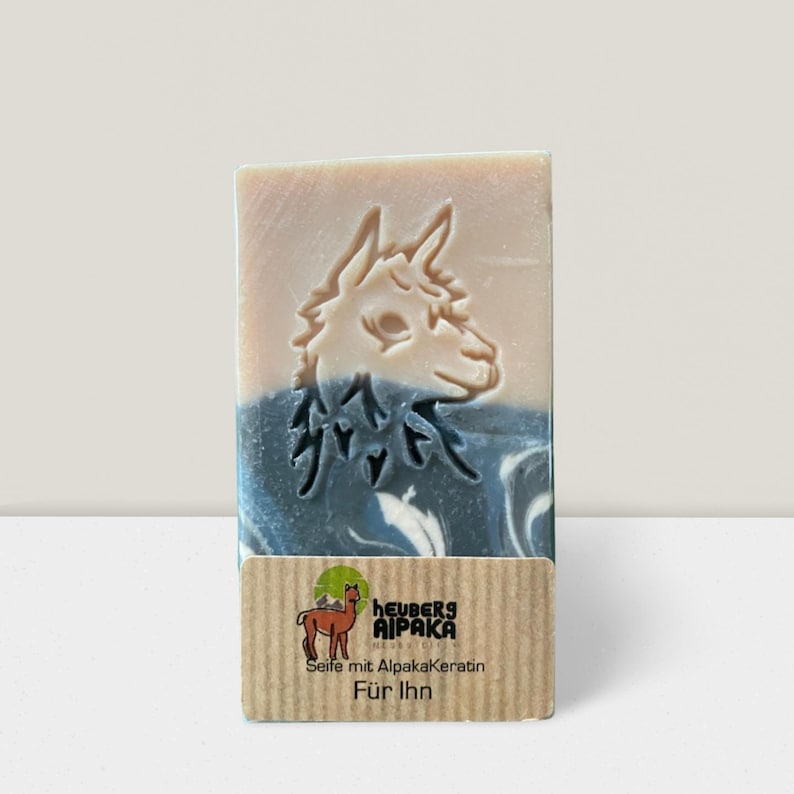 May include: A rectangular bar of soap with a carved alpaca design. The soap has a light beige top, a blue and white marbled center, and a brown label with text and a logo. The label reads "HEUBERS AIPAKA" and "Seife mit Alpakakeratin F&uuml;r Ihn."