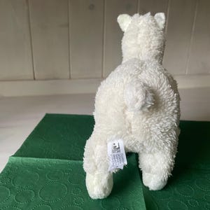 May include: A white plush polar bear toy, seen from behind, with a fluffy texture. The bear is placed on a green patterned surface. The toy has a small tail and a tag attached to its rear.
