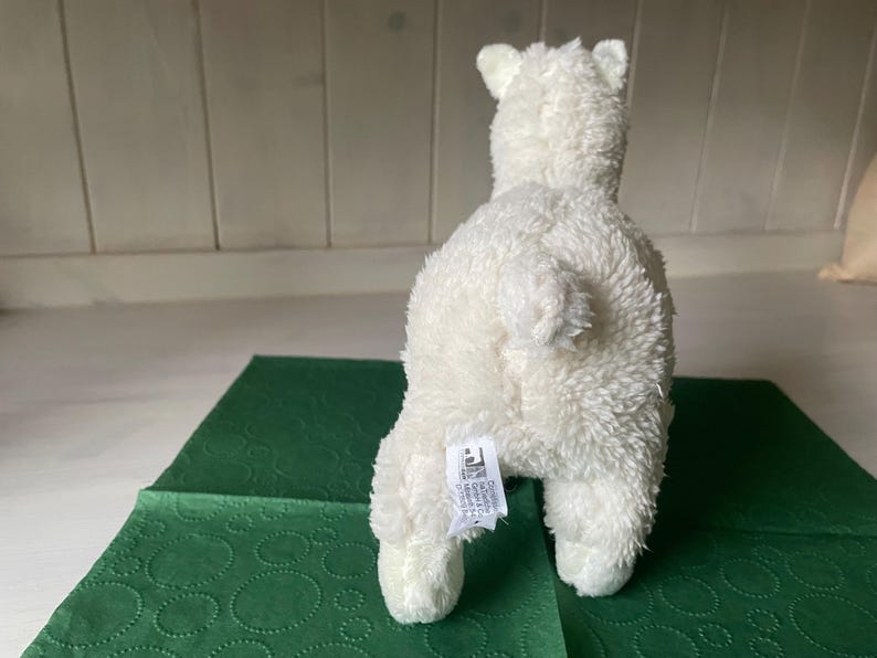 May include: A white plush polar bear toy, seen from behind, with a fluffy texture. The bear is placed on a green patterned surface. The toy has a small tail and a tag attached to its rear.