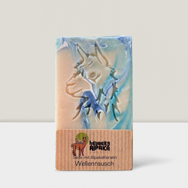 May include: A rectangular bar of soap with a carved alpaca design. The soap has a light beige base with blue and white swirls. A brown label at the bottom features the brand name and product details.