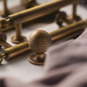 May include: Close-up of antique brass hardware, including a round knob with a grooved design and several matching bar pulls. The hardware has a warm, aged finish, suggesting a vintage or classic style. The items are arranged on a light surface.