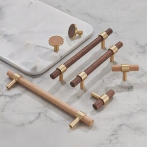 以下が含まれることがあります： 木製のキャビネット金具のコレクション。つまみと引き手が含まれています。明るい木材と濃い木材、真鍮色の金属アクセントを組み合わせています。大理石模様の表面に配置され、デザインと仕上げが際立っています。