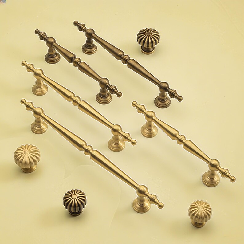 May include: A selection of antique brass cabinet handles and knobs. The image shows long bar pulls and round knobs with decorative detailing. The hardware is arranged on a light yellow surface. Ideal for furniture and home decor projects.
