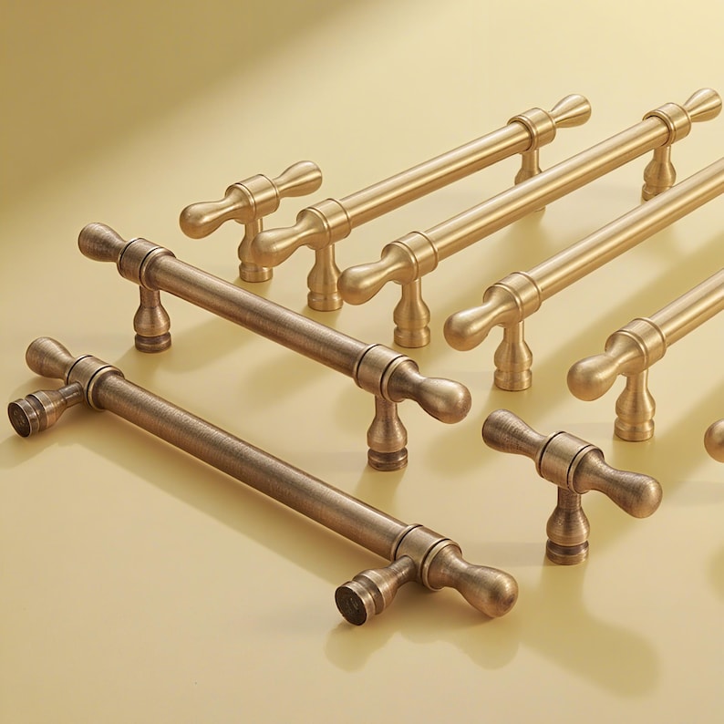 Op de afbeelding: Een verzameling bronzen en goudkleurige kastgrepen. De grepen hebben een cilindrische staaf met decoratieve uiteinden en zijn gerangschikt op een lichtgekleurd oppervlak. De hardware is ontworpen voor meubels en kasten.