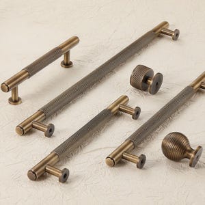 May include: A selection of antique brass cabinet hardware, including pull handles and knobs. The handles feature a ribbed design and are mounted on small posts. The knobs have a similar ribbed design. The hardware is arranged on a light-coloured surface.