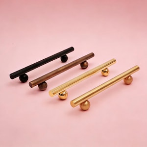 May include: Four cabinet pulls in a row, each with a cylindrical bar and a spherical base. The pulls are in black, bronze, and two shades of gold. The background is a solid pink.
