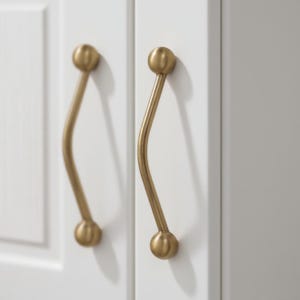 Może przedstawiać: Zbliżenie dwóch złotych uchwytów do szafek o zakrzywionym kształcie. Uchwyty są zamontowane na białych drzwiach szafki. Każdy uchwyt ma kulisty koniec i zakrzywiony, smukły korpus.