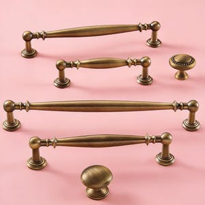 May include: An assortment of antique brass cabinet hardware, including four pull handles and two knobs. The handles vary in size, with a curved design and decorative detailing. The knobs are round with a similar finish. All pieces are displayed on a pink background.