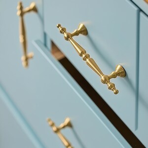 May include: Close-up of a pale blue cabinet with gold-coloured handles. The handles have an ornate design and are attached to the cabinet doors. The cabinet doors are partially open, revealing the interior.