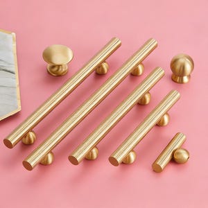Può includere: Una collezione di ferramenta per mobili color oro, tra cui maniglie a barra cilindriche e pomelli rotondi. Le maniglie variano in lunghezza e i pomelli hanno un design arrotondato. L'hardware è esposto su una superficie rosa, mostrando un'estetica moderna.
