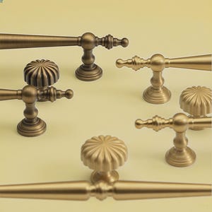 May include: A selection of antique brass cabinet handles and knobs. The handles are lever-style, and the knobs have a fluted, rounded design. The hardware is shown against a pale yellow background. The cabinet hardware is suitable for furniture.