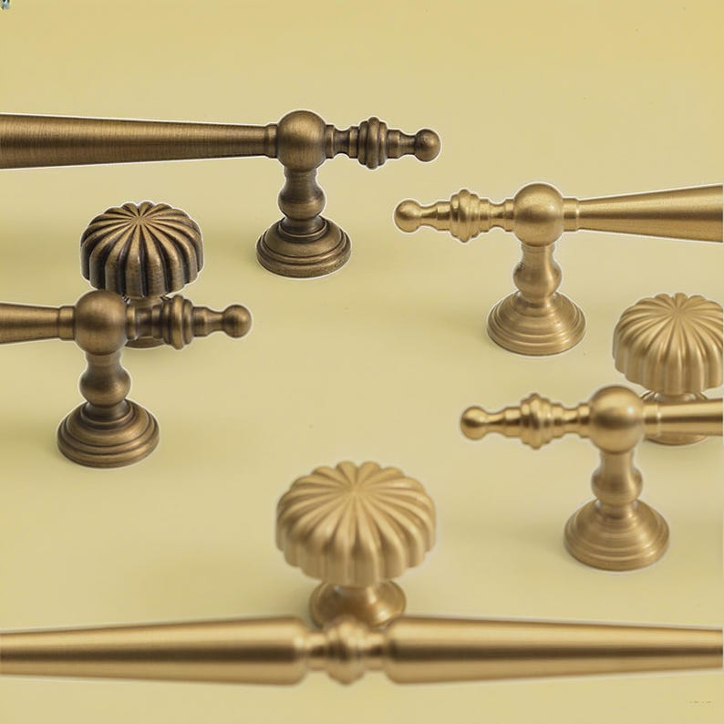 May include: A selection of antique brass cabinet handles and knobs. The handles are lever-style, and the knobs have a fluted, rounded design. The hardware is shown against a pale yellow background. The cabinet hardware is suitable for furniture.