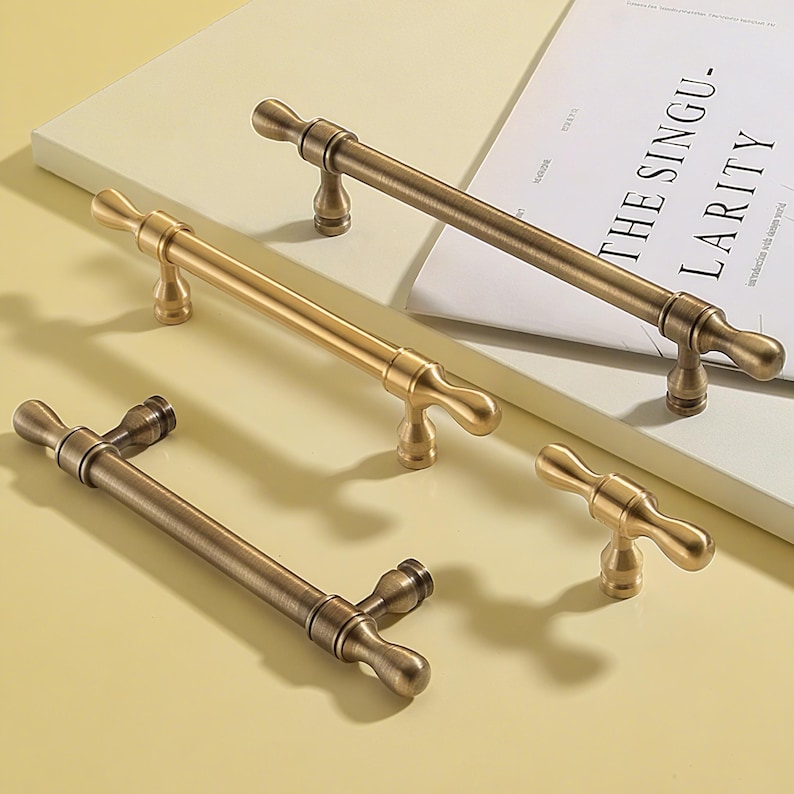 Op de afbeelding: Een assortiment antiek messing kastgrepen in verschillende maten. De grepen hebben een klassiek T-bar ontwerp met afgeronde uiteinden en een centrale, decoratieve steel. Sommige zijn donker bronskleurig, andere goudkleurig.