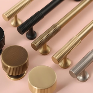 May include: Close-up of various cabinet hardware, including knobs and pulls. The knobs are black, bronze, and gold, with a ribbed texture. The pulls are gold, black, and silver, with a ribbed center section. All hardware has a modern design.