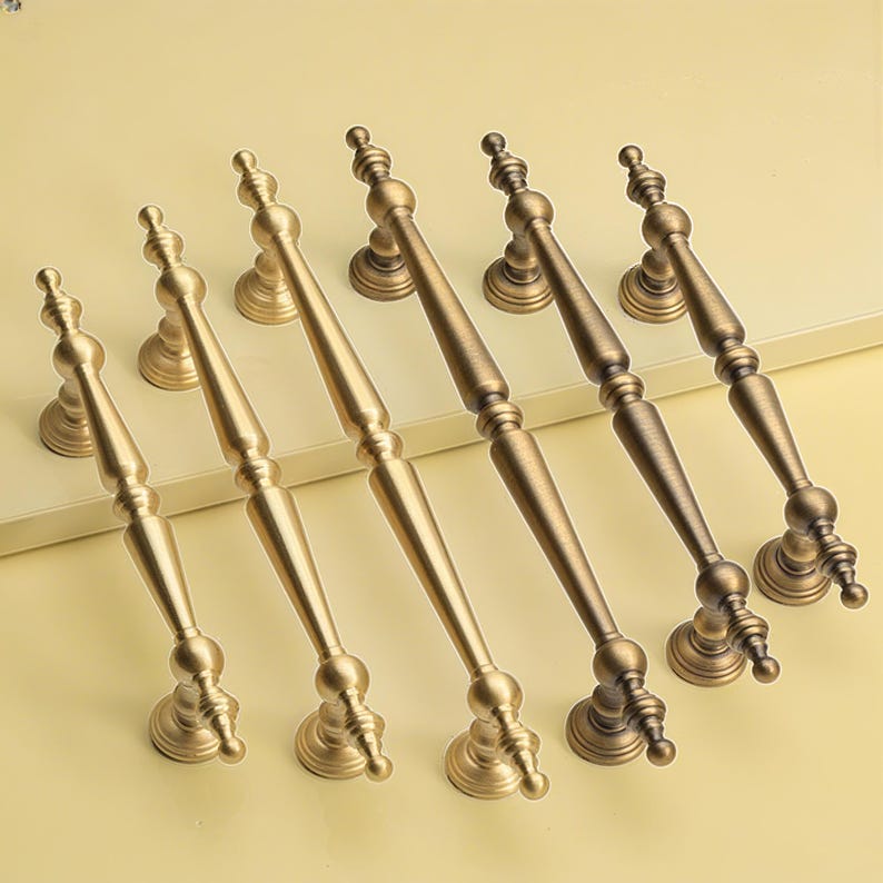 May include: Six antique-style cabinet handles in shades of gold and bronze. Each handle has an elongated design with a central grip and ornate end pieces. The handles are arranged on a light yellow surface.