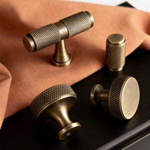 May include: A collection of antique brass cabinet hardware, including a T-bar handle, a cylindrical knob, and two mushroom-shaped knobs. Each piece features a textured, diamond-patterned surface and a warm, aged finish. The hardware is displayed on a black surface with a peach-colored fabric.