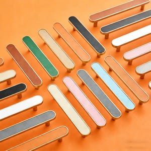May include: A selection of cabinet pulls in various colours, each with a gold-coloured frame. The pulls are in shades of brown, green, black, white, pink, and blue. They are arranged on an orange background.