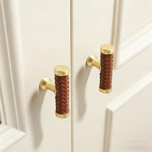 Könnte beinhalten: Zwei Schrankgriffe mit goldener Oberfläche und braunen Ledergeflecht-Details. Die zylindrischen Griffe haben eine goldene Kappe und einen goldenen Sockel und sind an einer weißen Schranktür montiert. Das Ledergeflecht verleiht Textur und Wärme.