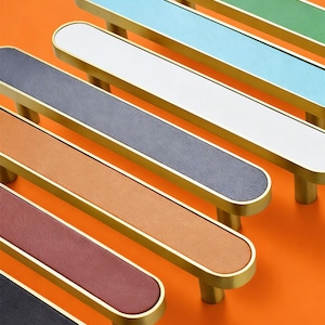 Puede incluir: Una colección de tiradores de armario en varios colores, incluyendo verde, azul, blanco, gris, marrón, rojo y negro. Cada tirador presenta un centro rectangular con un marco dorado y postes cilíndricos.