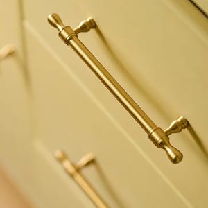 Op de afbeelding: Een goudkleurige kastgreep met een cilindrische stang en decoratieve uiteinden. De hardware is gemonteerd op een lichtgele kastdeur. De greep is een klassiek ontwerp voor keuken- of badkamerkasten.