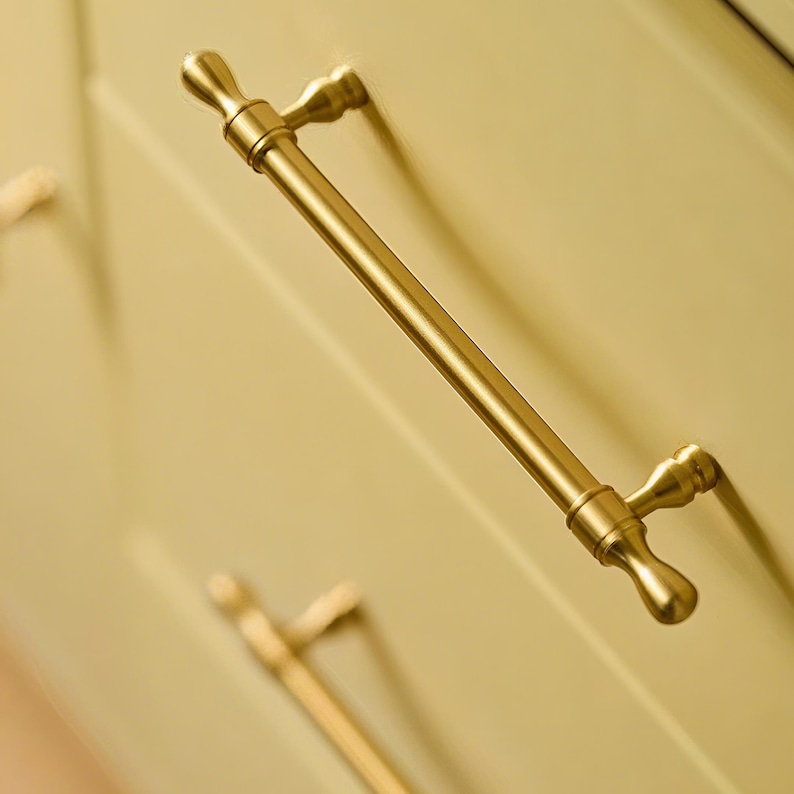 Op de afbeelding: Een goudkleurige kastgreep met een cilindrische stang en decoratieve uiteinden. De hardware is gemonteerd op een lichtgele kastdeur. De greep is een klassiek ontwerp voor keuken- of badkamerkasten.