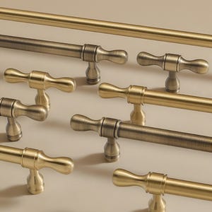 Op de afbeelding: Close-up van verschillende antieke messing kastgrepen en een bijpassende stanggreephandgreep. De grepen hebben een klassiek, elegant ontwerp met een gebogen handvat en een decoratieve basis. De afwerking varieert van helder goud tot donkerder, verouderd brons.