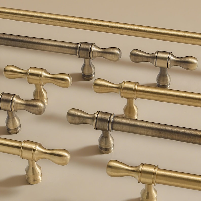 Op de afbeelding: Close-up van verschillende antieke messing kastgrepen en een bijpassende stanggreephandgreep. De grepen hebben een klassiek, elegant ontwerp met een gebogen handvat en een decoratieve basis. De afwerking varieert van helder goud tot donkerder, verouderd brons.