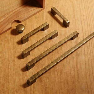 May include: A collection of antique brass cabinet hardware, including several rectangular bar pulls and a round knob. The hardware is arranged on a light wood surface, showcasing the aged finish and design of the pulls.