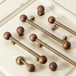 May include: A collection of cabinet hardware featuring wooden ball knobs and bronze-toned bar pulls. The knobs and pulls are arranged on a cream-colored surface, showcasing their design and finish. The hardware is designed for furniture and cabinetry.