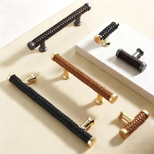 May include: A collection of decorative cabinet pulls with woven leather-like handles. The pulls come in various colors, including black, brown, and tan, with gold or dark metal accents. The handles are approximately 6 inches long.