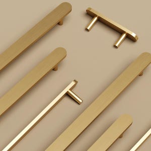 May include: A collection of gold-colored cabinet hardware, including long bar pulls and T-shaped knobs. The hardware has a brushed finish and a modern design, suitable for kitchen or bathroom cabinets. The hardware is shown against a neutral background.
