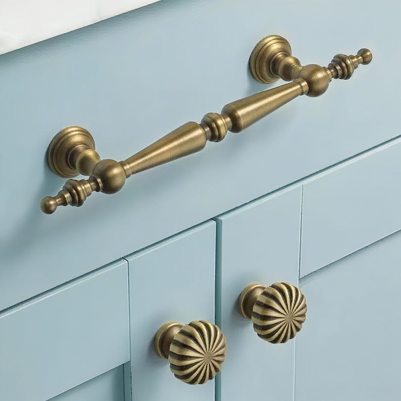 May include: Close-up of antique brass cabinet hardware. The image shows a long handle and two round knobs with a sunburst design. The hardware is mounted on a light blue cabinet.