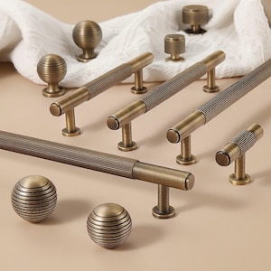 May include: An assortment of antique brass cabinet hardware, including ribbed bar pulls, round knobs, and spherical knobs. The hardware has a warm, aged finish, and is arranged on a neutral background with a white cloth.