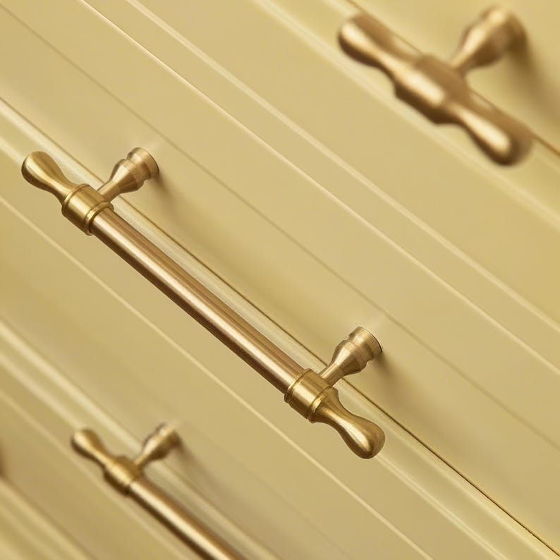 Op de afbeelding: Close-up van een cr&egrave;mekleurige kast met verschillende goudkleurige metalen handgrepen. De handgrepen zijn staafvormig met decoratieve uiteinden. De kastdeuren hebben een groefontwerp en de handgrepen zijn diagonaal gemonteerd.