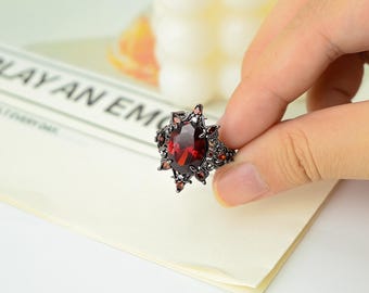 May include: A dark, ornate ring featuring a large, oval-cut, deep red gemstone as the centerpiece. The ring has a black metal setting with smaller red accent stones and a starburst design around the main stone. The ring is held by a hand.