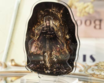 May include: A clear acrylic stand featuring a detailed illustration of a figure in dark attire, holding a sword. The artwork has a fantasy theme with gold and brown accents, set against a dark background. The stand is on a clear base.