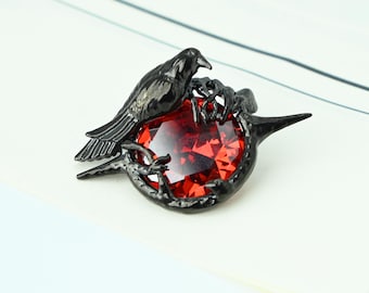 May include: A black raven perched on a red gemstone, set in a dark metal frame. The raven is detailed with visible feathers, and the gemstone has a faceted appearance. The jewelry piece is likely a brooch or pendant.