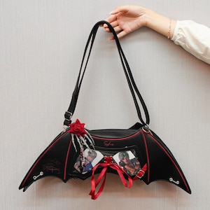 Może przedstawiać: Czarna torebka w kształcie skrzydła nietoperza z czerwonymi wykończeniami i akcentami. Torba posiada długi pasek na ramię oraz ozdobny czerwony kwiat róży i zawieszkę w kształcie dłoni szkieletu. Dwa zdjęcia są przymocowane czerwoną wstążką.