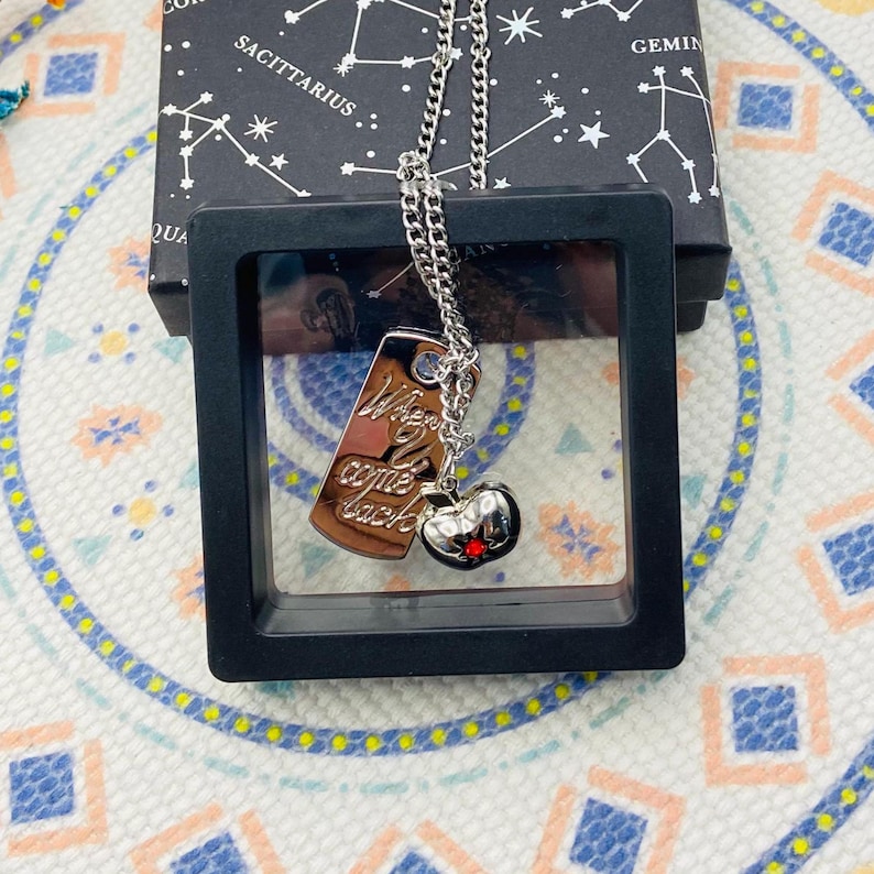 Puede incluir: Un collar de plata con un dije de placa de plata que dice "When you come back" y un dije de coraz&oacute;n rojo con una corona de plata. El collar est&aacute; en una caja negra con un patr&oacute;n de estrellas.