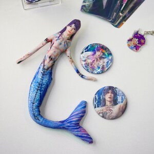 May include: A fabric mermaid doll with a blue and purple tail, and a light skin tone. Two round buttons and a keychain with a similar design are also present. The mermaid has long purple hair and is lying on a white surface.