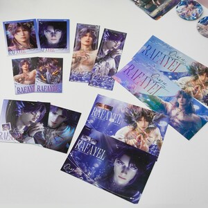 May include: A collection of printed cards and bookmarks featuring a fantasy character with long purple hair. The cards display the name "RAFAYEL" and other text in a stylized font. The color palette includes blues, purples, and whites.