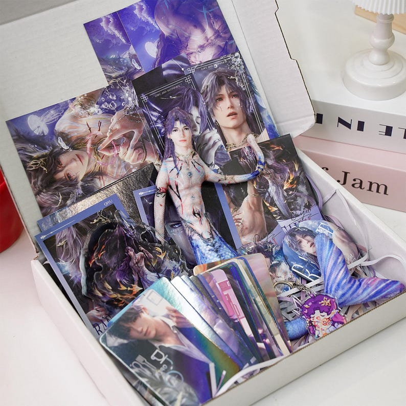 May include: A white box filled with fantasy-themed merchandise. The box contains various items, including art prints, trading cards, and a small figurine. The prints and cards feature illustrations of male characters with long hair and fantasy attire. The figurine is a mermaid-like character.