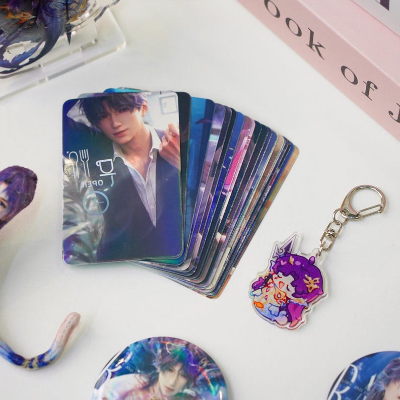 May include: A stack of glossy trading cards featuring a person in a suit, with a fork and knife icon. A clear acrylic keychain with a cartoon character and a silver keyring. Other merchandise is visible.