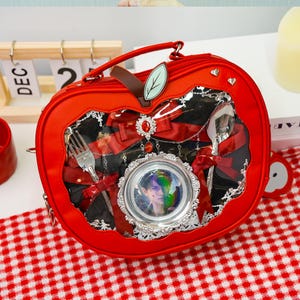 Może przedstawiać: Czerwona torebka w kształcie jabłka z przezroczystym oknem, odsłaniającym dekoracyjne sztućce, czerwone wstążki i oprawiony portret. Torebka ma uchwyt, akcent w kształcie liścia i zapięcie na zamek błyskawiczny. Torebka leży na czerwono-białej kratowanej powierzchni.