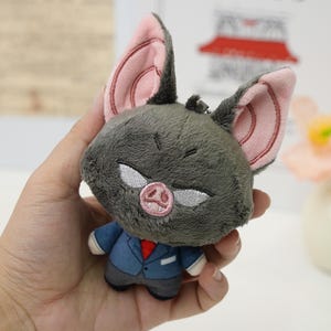 May include: A plush bat toy with a gray body, large pink-lined ears, and a small snout. The bat is wearing a blue suit jacket, gray pants, and a red tie. The toy is held in a hand.